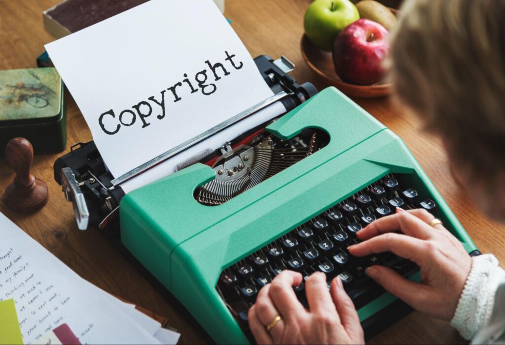 Фотография рук, печатающих на винтажной зеленой печатной машинке со словом "Copyright" на листе бумаги, рядом лежат яблоки и документы